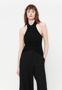 Kvinne med rødt hår iført en ermeløs svart halterneck-top og vide sorte bukser, stående mot en enkel hvit bakgrunn.