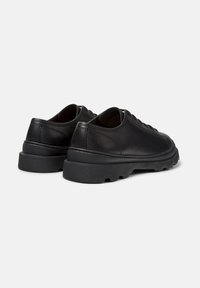 Un paio di scarpe in pelle nera con lacci e suole in gomma spessa e testurizzata, viste da dietro su uno sfondo bianco.
