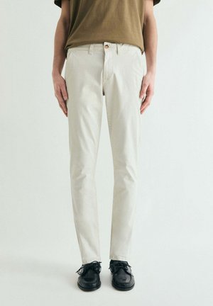 Persona con pantalones beige de corte recto, camisa marrón metida por dentro y zapatos negros con cordones, de pie contra un fondo claro y liso.