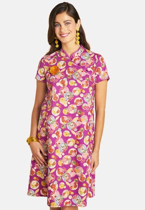 Woman wearing a purple floral patterned dress with short sleeves, yellow flower brooch, yellow earrings, and gold bangles, smiling at the camera.