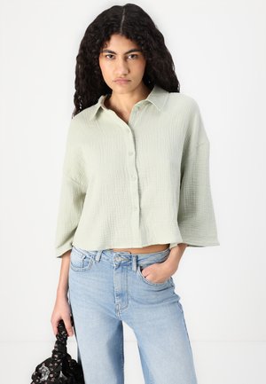 Light green, wrinkled fabric button-up shirt with a relaxed fit and three-quarter sleeves, paired with light blue jeans and a black bag.