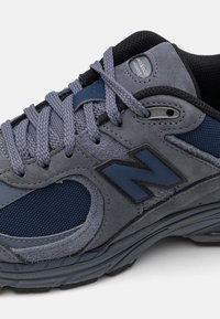 Gray and navy New Balance sneaker with textured suede and mesh, thick sole, and matching gray laces tied in a bow.