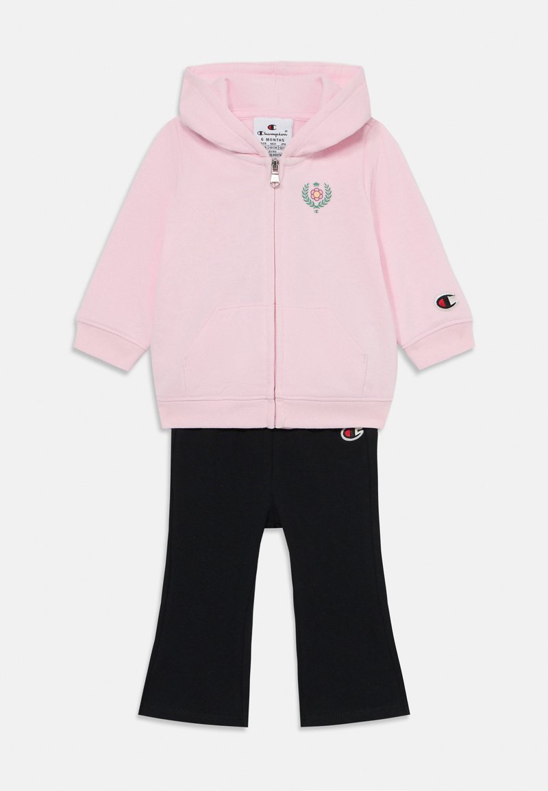 Felpa rosa con zip e logo floreale, abbinata a pantaloni neri con un patch del logo. Entrambi sono realizzati in tessuto morbido con un design casual.