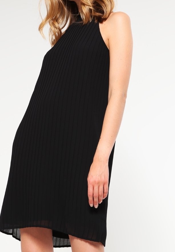 Femme portant une robe noire côtelée sans manches à col montant, debout devant un fond blanc uni.