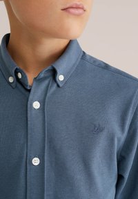 Zils krekls ar pogu aizdari, teksturēta auduma, ar izšūtu logotipu uz krūts, baltas pogas un pielāgota piegriezuma.