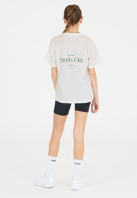 T-shirt blanche oversize avec le texte vert "Les Arcs Sports Club Luxury Athleisure Wear" au dos, assortie à un short noir et des baskets blanches.