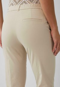 Pantalon beige avec une texture lisse, coupe fuselée et une poche arrière. Ceinture élastique pour plus de confort. Design minimaliste avec des lignes épurées.