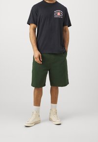 Czarny t-shirt z nadrukiem "Nike Hoops", zielone szorty i kremowe wysokie trampki. Widoczne białe skarpetki, luźny krój, lekka bawełniana tkanina.