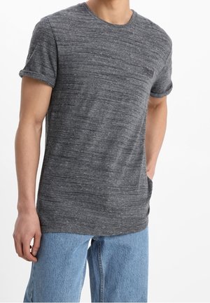 Mężczyzna ubrany w krótkim rękawem ciemnoszary melanżowy t-shirt i jasnoniebieskie dżinsy, stojący z jedną ręką w kieszeni na białym tle.