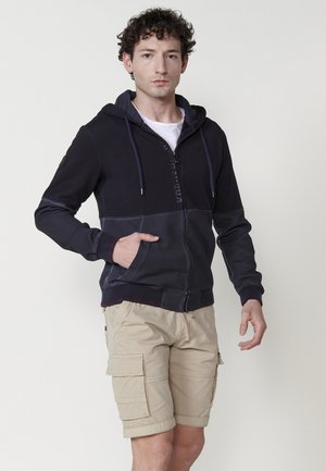 Felpa con cappuccio e zip di colore blu scuro e navy, in misto cotone, con pannelli a contrasto e tasca a marsupi. Abbinata a pantaloni cargo beige.