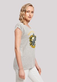 Grå t-skjorte med korte ermer som har et Hufflepuff-emblem i gult og svart, løst tilpasset, med en myk tekstur og rund halslinning.