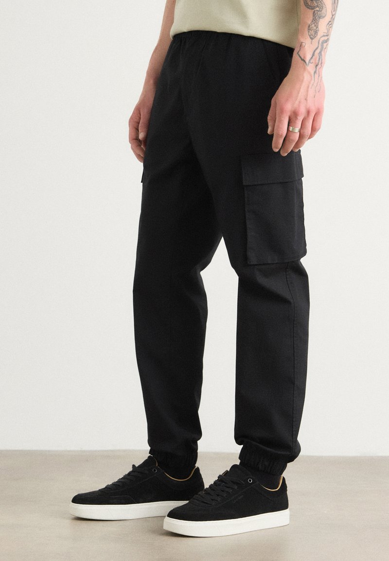 Zwarte cargobroek met elastische tailleband en boorden, met twee zijzakken. Gekleed met zwarte suède sneakers en een licht shirt.