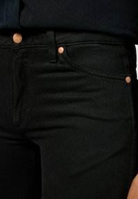 Gros plan sur un jean en denim noir avec des rivets et un bouton en cuivre, montrant les détails de la poche avant et de la ceinture.