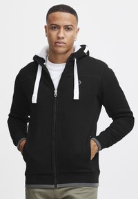 Crna zip-up kapuljača s kapuljačom obložnom bijelom bojom, sivim manžetama i džepovima. Mekani materijal s logotipom na prsima i kontrastnim detaljima u šivanju.