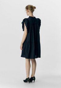 Schwarzes Kleid mit kurzen, Rüschensäumen, gestuftem Design und geraffter Taille. Das Material wirkt leicht und hat eine glatte Textur. Schwarze High Heels.