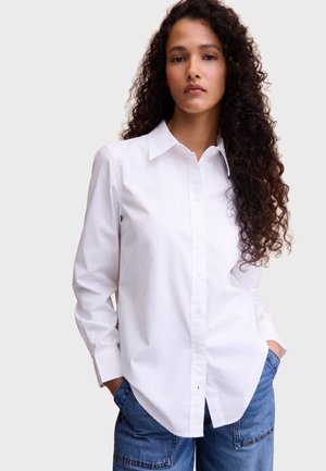 Donna con capelli ricci che indossa una camicia bianca con bottoni e jeans blu con le mani in tasca, in piedi davanti a uno sfondo chiaro e semplice.