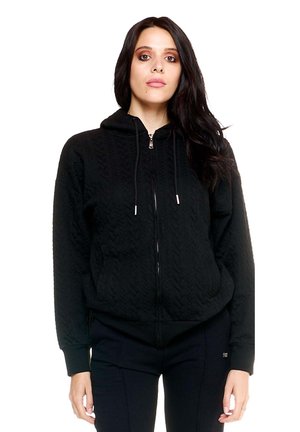 Une femme aux longs cheveux foncés porte un sweat à capuche noir matelassé et zippé, ainsi qu’un pantalon foncé, sur un fond blanc uni.