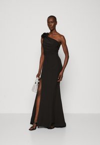 Rochie neagră cu un umăr, decorată cu o floare, având o fanta înaltă și un design lung până la podea. Material neted, texturat, asociat cu o geantă texturată.