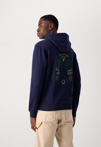 Haut à capuche bleu marine avec un cordon de serrage et des poignets côtelés. Présente un design graphique au dos avec des éléments sur le thème du tennis en vert et or.