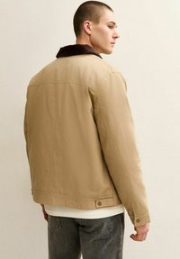 Ung man som står i profil, iklädd en beige jacka med mörkrandig krage, vit skjorta och mörkgrå jeans mot en ljus vägg.