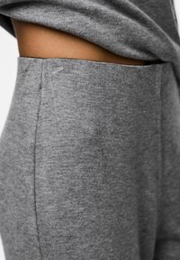 Tuvumā redzama persona, kas valkā augstās vidukļa pelēkās bikses un pelēko topu, kas atklāj vēderu, izceļot auduma tekstūru un sānu šuvju detaļas.