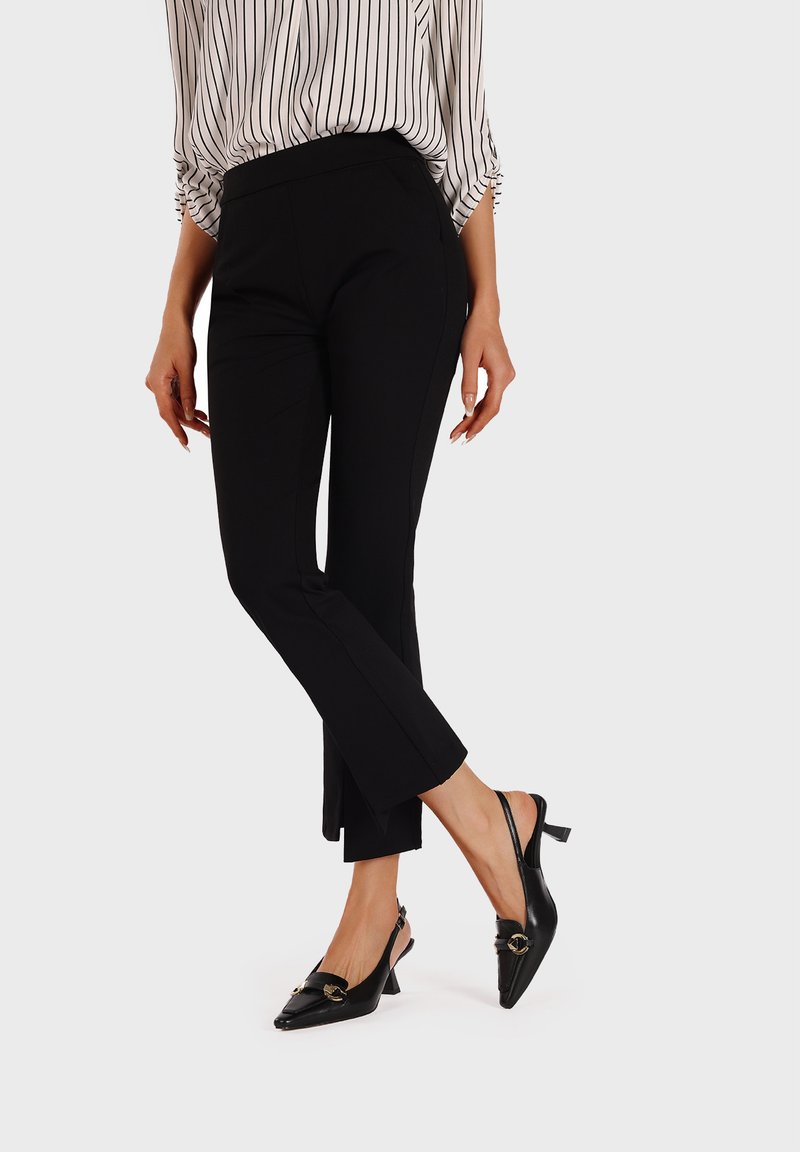 Pantaloni neri su misura a gamba stretta, con una texture liscia e design alla caviglia, abbinati a décolleté neri con tacco a scomparsa e dettaglio con fibbia.