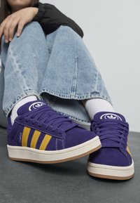 Oseba v vijoličnih supergah Adidas Campus z rumenimi črtami, svetlih modrih kavbojkah in belih nogavicah sedi na sivi tleh.