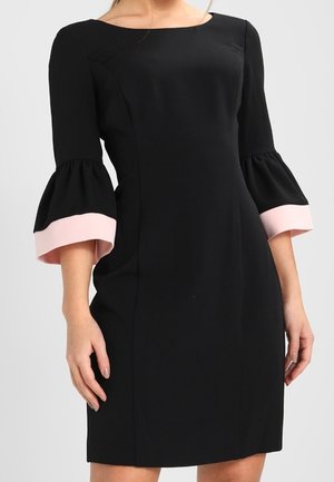 Sort knælængde kjole med treskåret klokkeærmer med lyserøde manchetter, båret af en person med hænderne afslappet ned langs siderne.