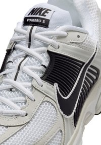 Nike Vomero 5 športni copati imajo belo mrežasto zgornji del s črnimi detajli, teksturiranimi prekrivnimi sloji in belimi vezalkami, zasnovani za zračnost in podporo.