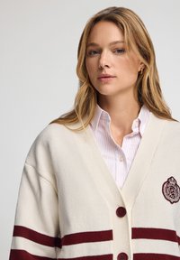 Krämfärgad cardigan med vinröda ränder, stickad textur och en logotyp i vintage-stil. Har knappar och är lagd över en rosa randig skjorta.