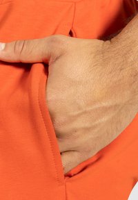 Orange tygficka med en hand insatt; slät textur och fint sydda kanter är synliga. En subtil hudton kontrasterar mot tyget.