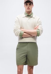 Νέος άνδρας που φοράει μπεζ πλεκτό πουλόβερ πάνω από πράσινο καρό πουκάμισο, πράσινα σορτς και καφέ καπέλο, στέκεται με σταυρωμένα τα χέρια μπροστά σε λευκό φόντο.