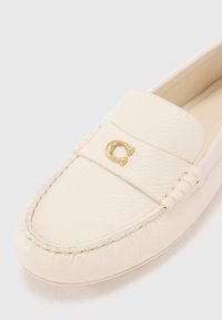 Valge nahast loafers, millel on õmmeldud disain, ümmargune nina ja kuldne logo aksent vampis. Sile tekstuur kerge läikega.