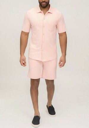 Homme portant une chemise à manches courtes rose clair à boutons et un short assorti, accompagné de chaussures noires à enfiler, sur un fond uni.