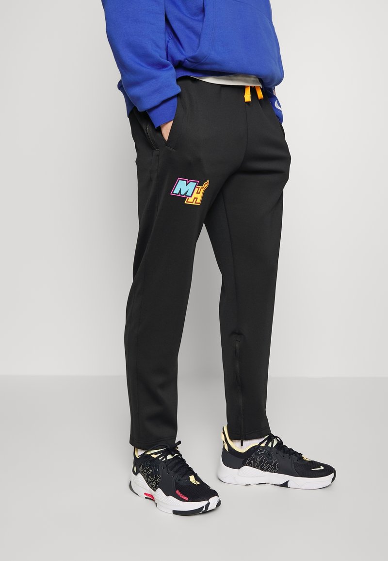 Personne portant un pantalon de survêtement noir avec un logo "MH" coloré et des chaussures de sport noires à semelles blanches, debout les mains dans les poches.