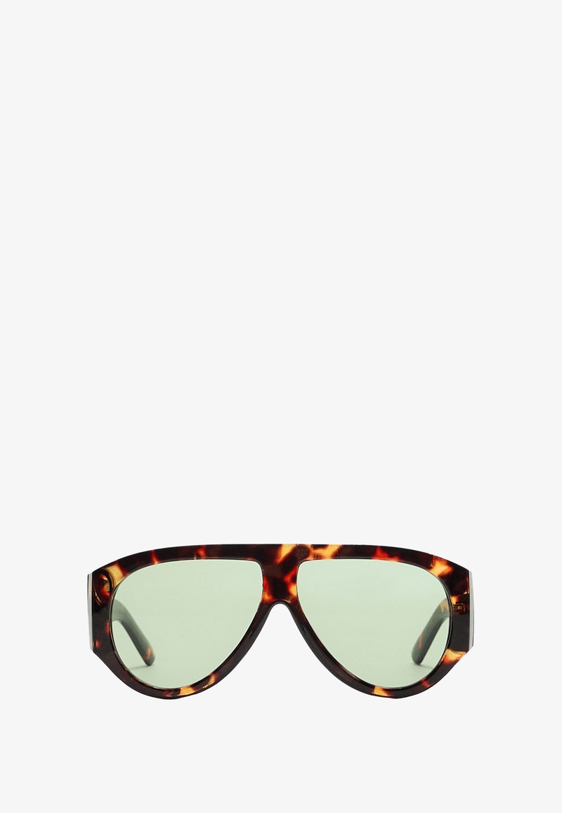 Sluneční brýle ve stylu želvoviny se zelenými skly, ostrým designem, širokými obrubami a výrazným obočím. Texturovaný povrch dodává hloubku.