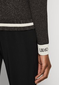 Maglione nero scintillante con una striscia bianca all'orlo e ai polsini, con un logo sul polsino. Tessuto liscio, design aderente.
