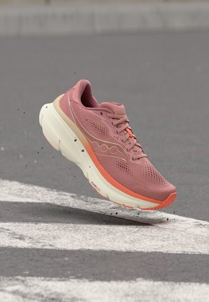 Roza in oranžen tekaški čevelj v zraku nad asfaltom z belimi cestnimi oznakami, prikazuje podplat in podrobnosti bočne znamke.