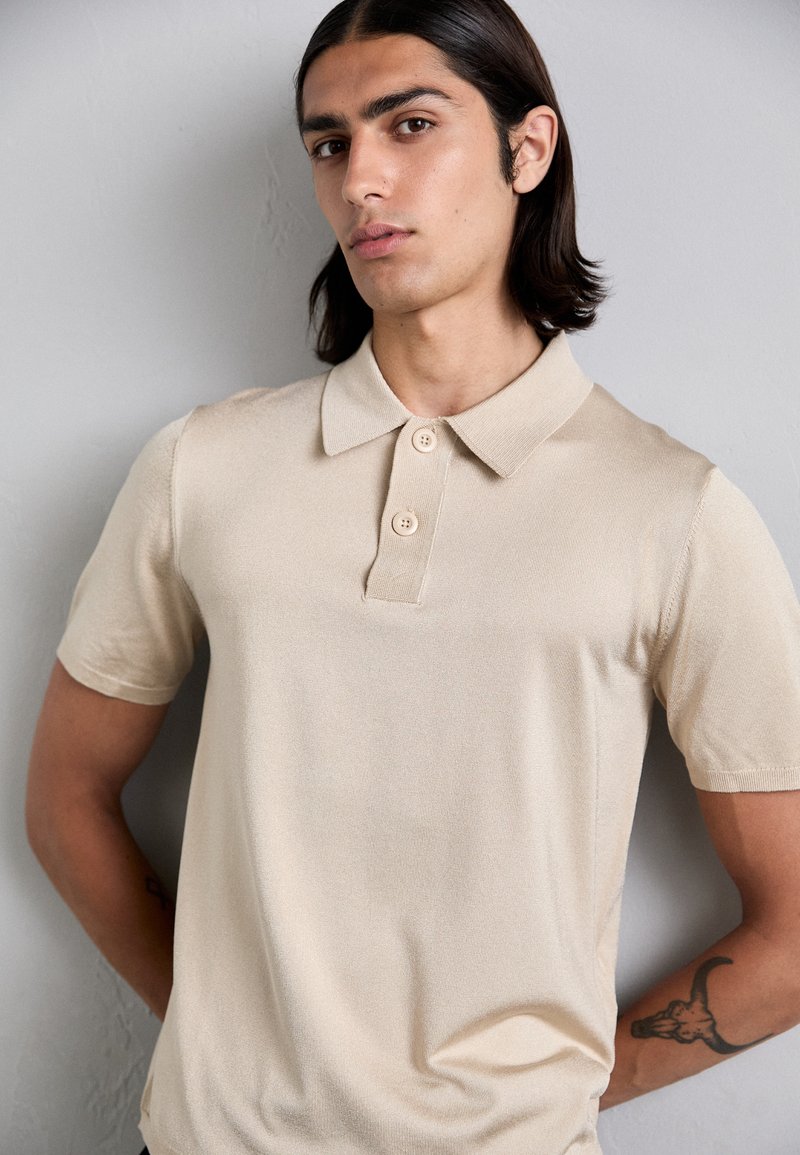 Beige polo skjorta i strukturerat tyg med korta ärmar, vikbar krage och två-knappars knäppning. Subtil lyster och figurpassande design.