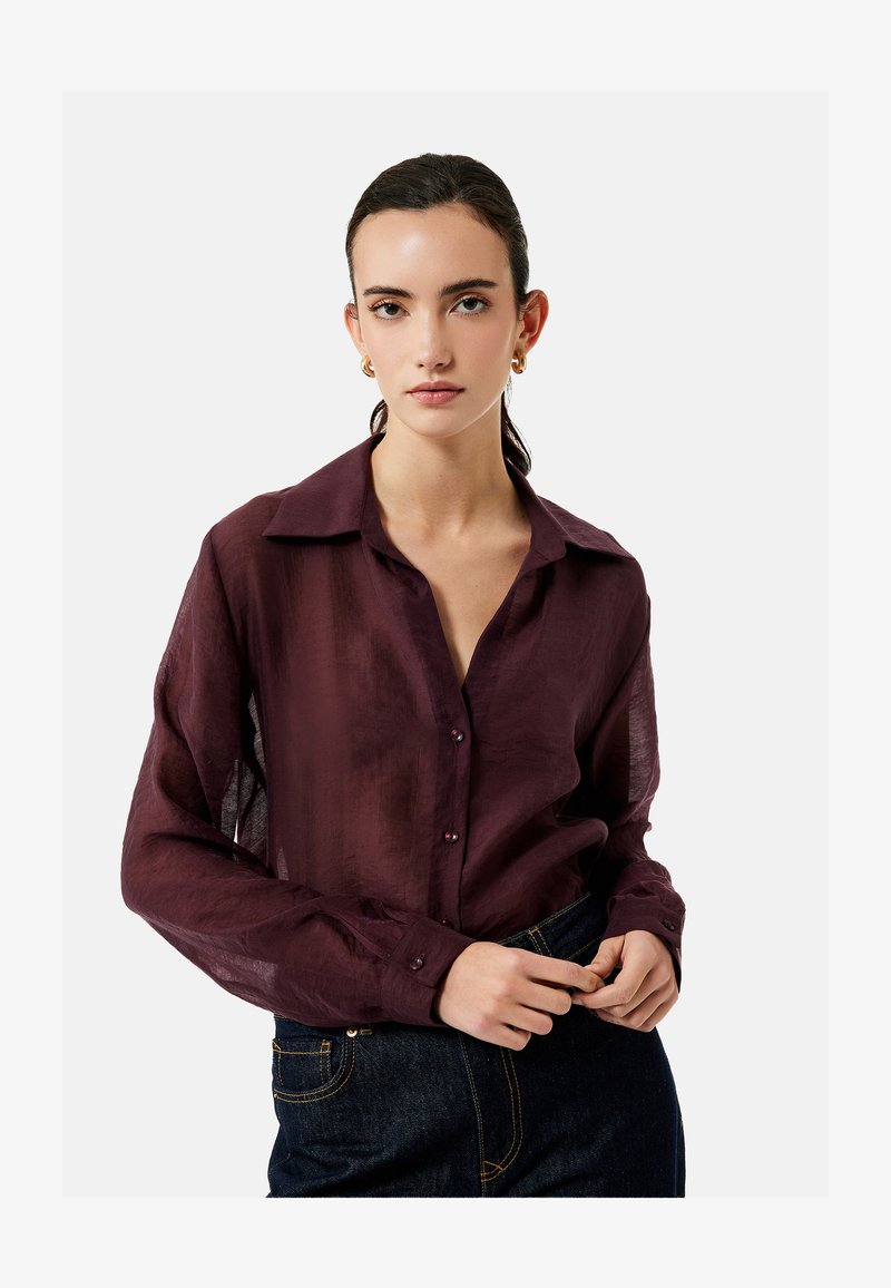 Mujer joven que lleva una blusa burdeos transparente con botones y vaqueros oscuros, con pendientes de aro dorados, de pie frente a un fondo liso.