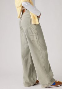 Άτομο  που φοράει μπεζ φαρδύ παντελόνι με τσέπη cargo, λευκό μπλουζάκι με μακριά μανίκια, κίτρινο πλεκτό πουλόβερ περασμένο στους ώμους και καφέ αθλητικά παπούτσια.