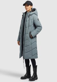 Padded long coat in light gray with a hood, featuring a quilted pattern, two side pockets, and a front zipper. Paired with black ankle boots.