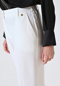 Weiße, plissierte Hose mit einem figurbetonten Bund, metallischem Akzent und glatter Stoffstruktur, kombiniert mit einem schwarzen Langarmtop.