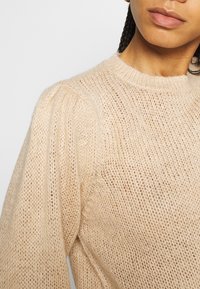 Vaaleanbeige neulepaita, jossa on kuvioitu, avoin kudos, puhvimaiset hihat ja klassinen pyöreä pääntie. Pehmeä kangas ja rento istuvuus.