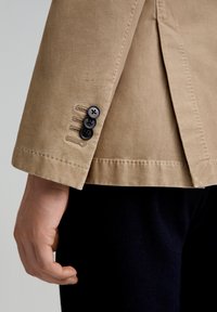 Blazer de algodón marrón claro con mangas y tres botones negros, costuras visibles y un corte limpio en el puño, combinado con pantalones de tela oscura.