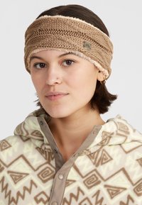 Knit headband in tan with a soft, plush lining. Features a textured pattern and a small metal brand tag on the side.