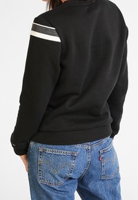 Sudadera negra con rayas grises y blancas en los hombros, combinada con jeans de mezclilla azules. Logo de Levi's visible en los bolsillos traseros.
