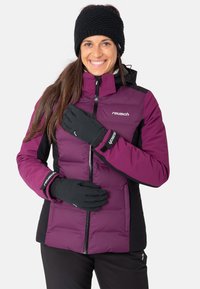 Lila Isolierjacke mit schwarzen Akzenten, Reißverschluss und Logo. Modell trägt schwarze Handschuhe und eine gestrickte schwarze Mütze.