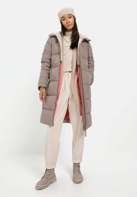 Lang grijze pufferjas met een capuchon, voorzien van een roze voering, gecombineerd met een lichtbeige trui en crèmekleurige broek. Grijze veterschoenen maken de look compleet.