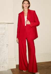 Ensemble de blazer cintré rouge et de pantalons à jambes larges en tissu texturé. Le blazer présente un col cranté et deux poches.